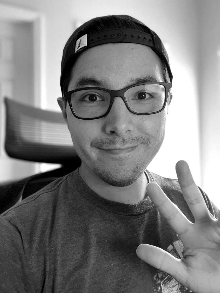 Jesse Ruiz wearing a dark cap and glasses, raising one hand in a wave while seated at a desk chair.